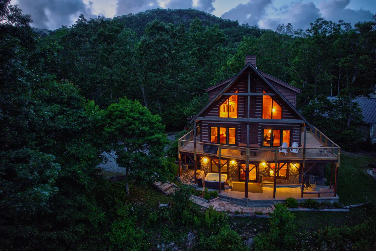 A warmly lit log cabin set among trees, with glowing windows and a bubbling outdoor hot tub. The spacious deck invites groups to gather under the open mountain sky, and the natural surroundings offer privacy and relaxation.