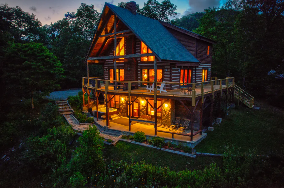 The hero image showcases a charming log cabin nestled among lush greenery, with a spacious deck adorned with inviting seating and glowing warm lights. The mountain setting at 3900' is evident, with expansive views visible from the deck, creating a cozy and welcoming atmosphere for gatherings, relaxation, or soaking in the scenery.