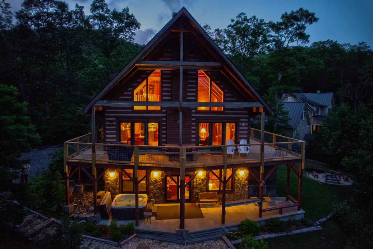 A warmly lit log cabin sits nestled among lush greenery, with an inviting outdoor hot tub and a spacious deck featuring comfortable seating. The setting exudes tranquility and privacy, with views of the surrounding woods and the promise of stargazing or sunset watching from the deck.