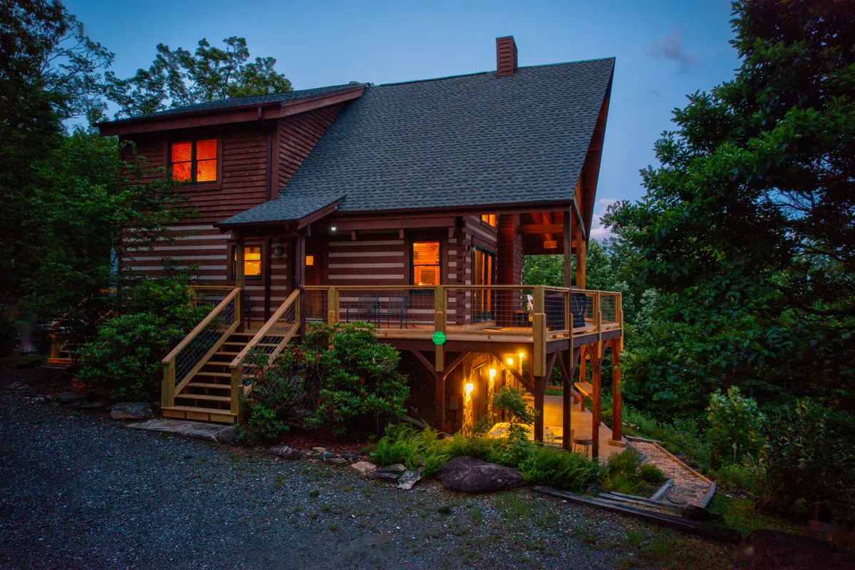 A glowing wooden deck illuminated by soft porch lights, with large windows revealing a welcoming, warm interior. The surrounding greenery and seating area evoke the ideal setting for barbecues, evening chats, and stargazing.