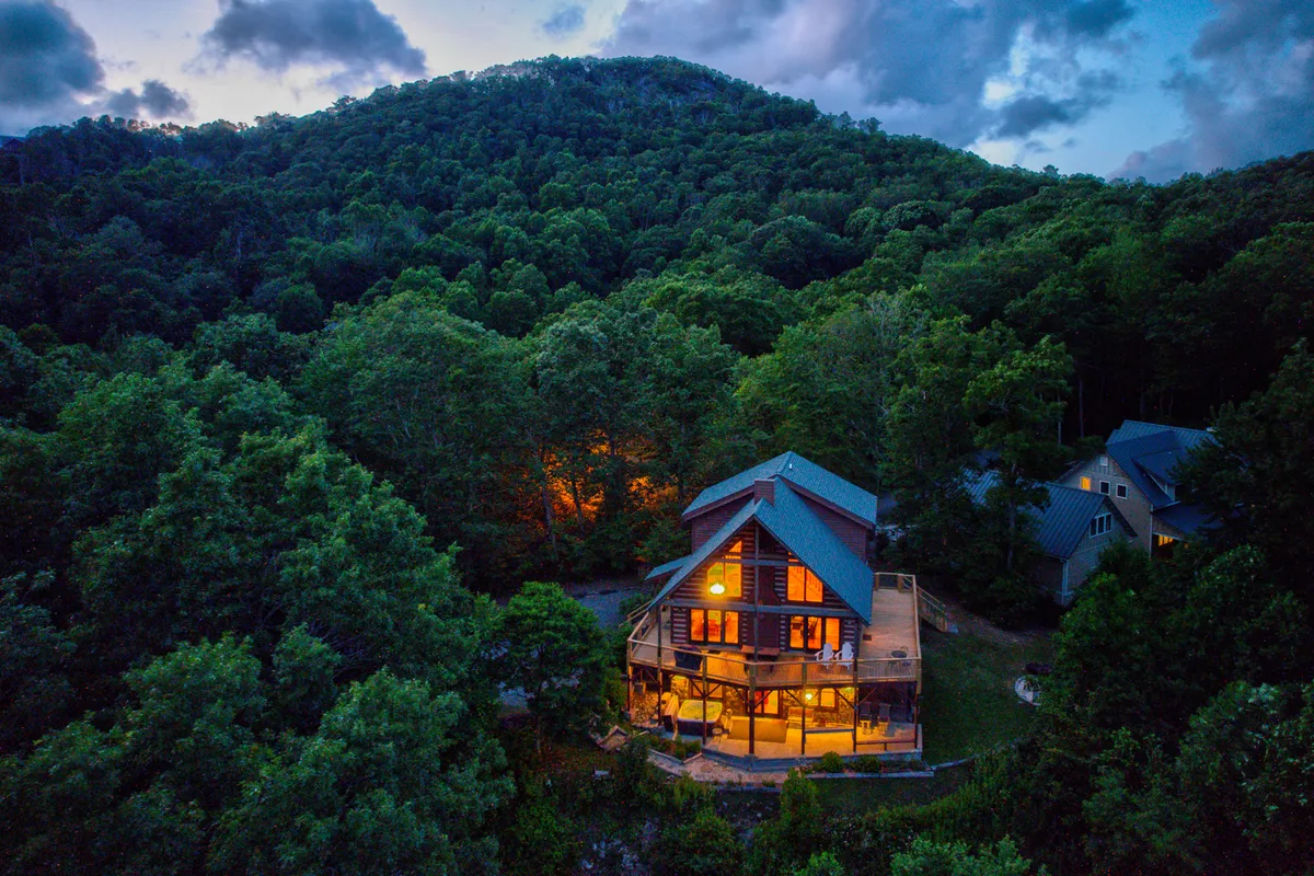A classic wooden cabin set against a breathtaking mountain landscape, featuring an expansive outdoor deck. The scene exudes a welcoming spirit and highlights the proximity to nature and space for larger group activities.