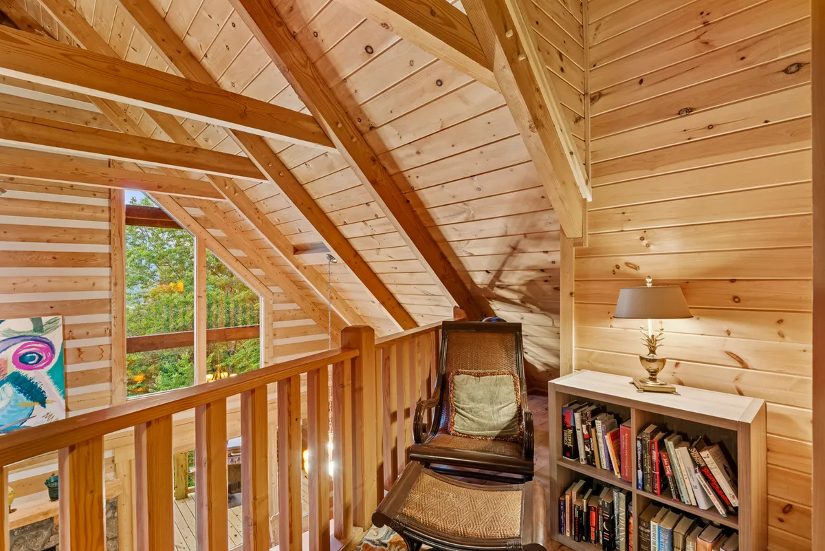 A tranquil loft space with wooden beams and large windows, featuring a cozy reading nook and a bookshelf. The nook overlooks scenic outdoor views, offering a peaceful spot for quiet reflection or enjoying a good book.