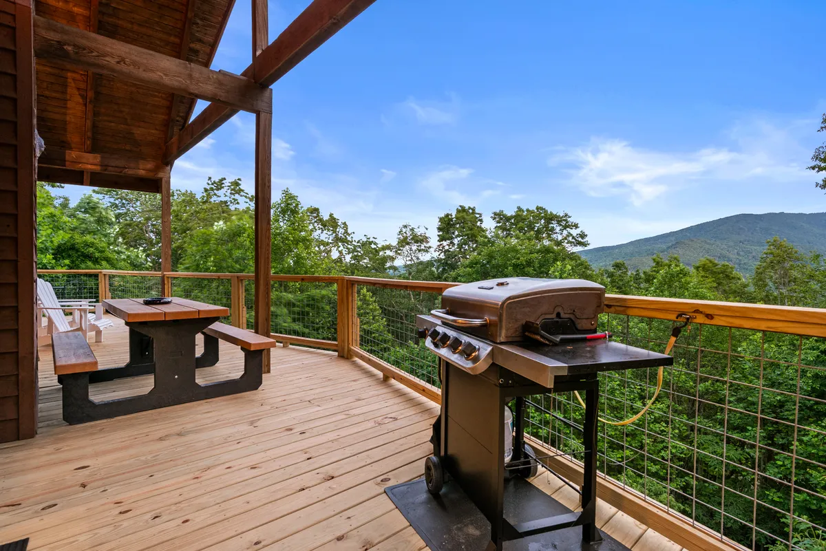 This image features a deck area with a grill and picnic table set against a sweeping mountain view. The scene suggests a casual, inviting atmosphere ideal for outdoor dining and barbecuing while soaking in the natural surroundings.