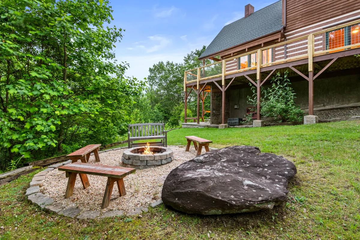 A cozy fire pit area is surrounded by ample seating and lush greenery, with a large rock feature adding to the natural ambiance. The setup is ideal for intimate gatherings, storytelling, and enjoying the outdoors after dark.