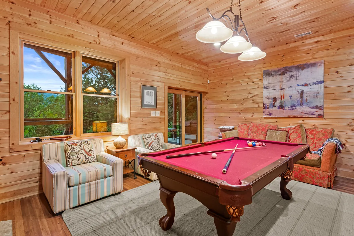 This image showcases a welcoming den featuring a pool table, striped armchair, floral sofa, and large windows that let in plenty of natural light. The setup evokes a casual, fun, and social atmosphere ideal for game nights and family gatherings.