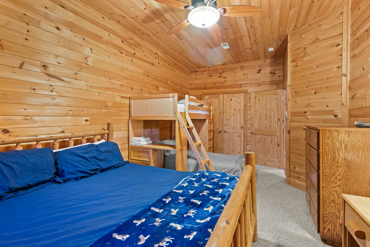 A spacious bedroom with warm wooden walls features a bunk bed and a double bed, complemented by a dresser and ceiling fan. The space’s relaxed ambiance suggests a perfect setting for family sleepovers, storytelling, or playing games together.