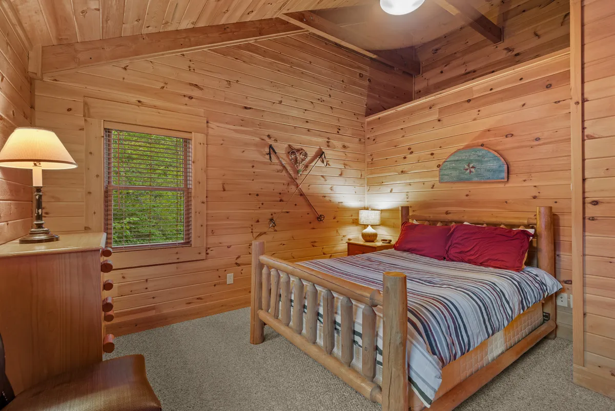 A warmly lit bedroom with a rustic log bed, striped bedding, and a nightstand lamp. Wooden walls and playful decor—including a decorative heart and surfboard—add a welcoming, personal touch. The window implies enjoyable views and a connection to nature.