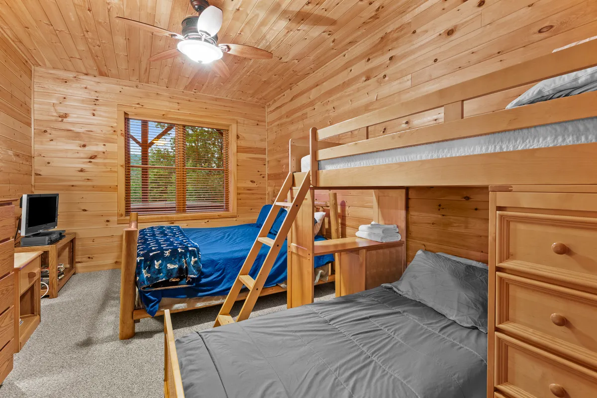 This image focuses on a bedroom with a bunk bed and a single bed, a dresser, and a TV. The window brings in morning sunlight, and the space looks inviting and perfect for children or groups seeking comfort and entertainment.