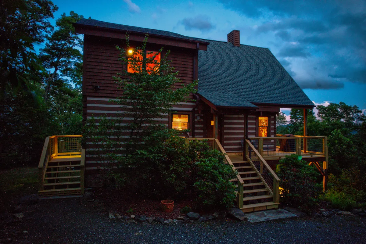 An idyllic view of a log cabin at dusk, with glowing windows hinting at warmth inside, a wooden deck for evening gatherings, and a backdrop of natural greenery. The scene evokes serenity, seclusion, and the allure of evening stargazing or sunset watching.