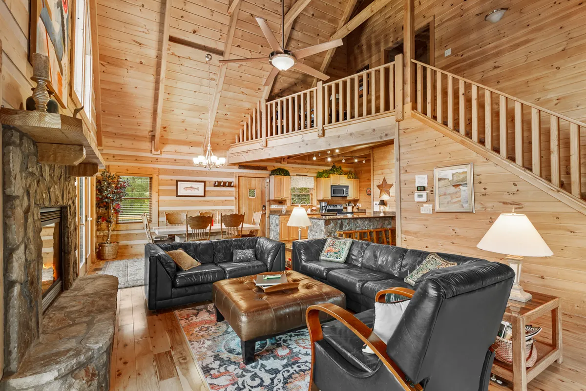 This image features a spacious open-plan great room with leather sofas, a striking stone fireplace, visible wooden beams, a dining area, and a staircase. The ambiance is relaxed and cozy, making it ideal for family gatherings and socializing. The fireplace and seating arrangement are central, conveying warmth and togetherness.